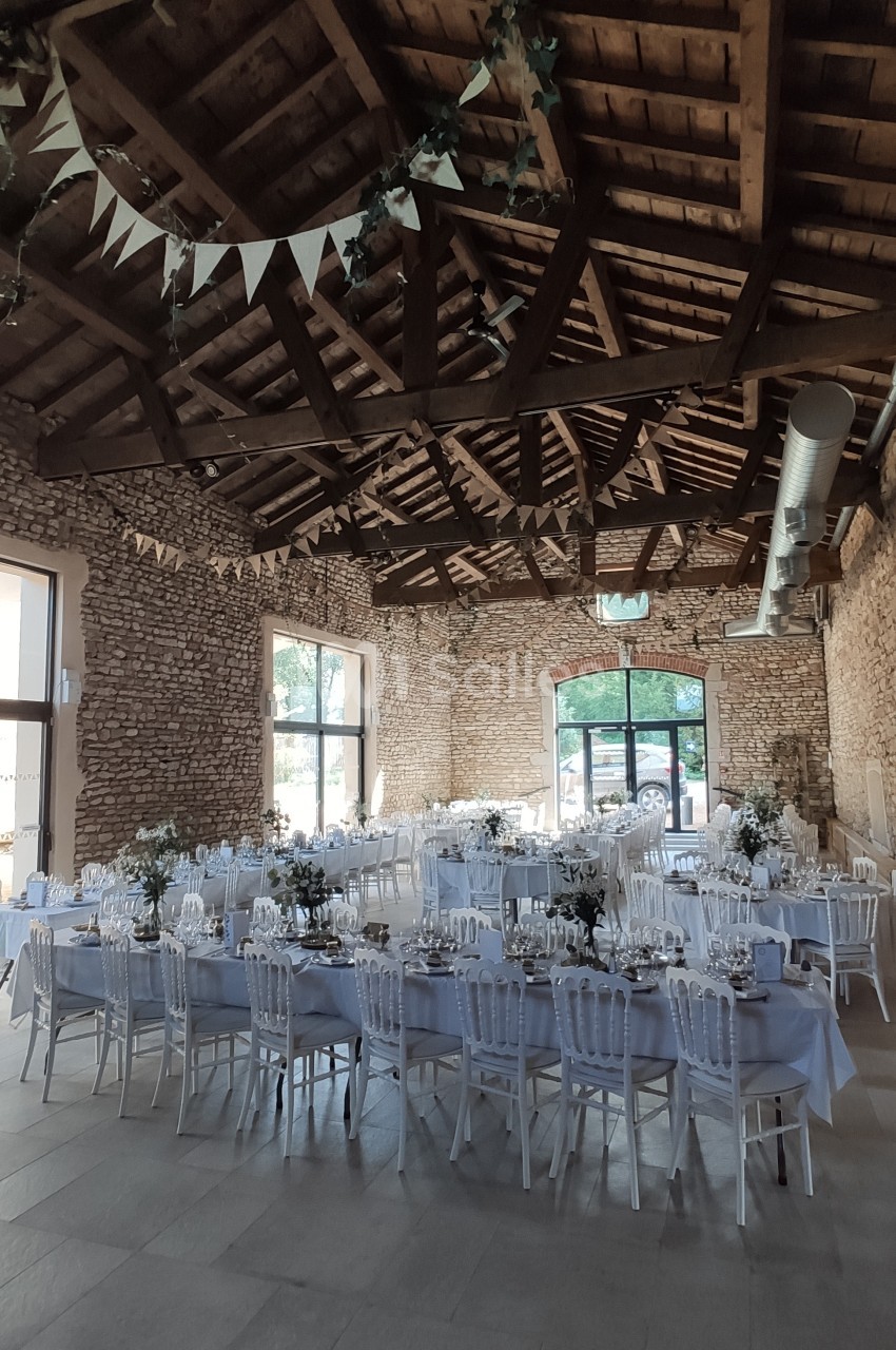 Salle de réception en pierre avec tables dressées, chaises blanches et décoration florale sous un plafond en bois.