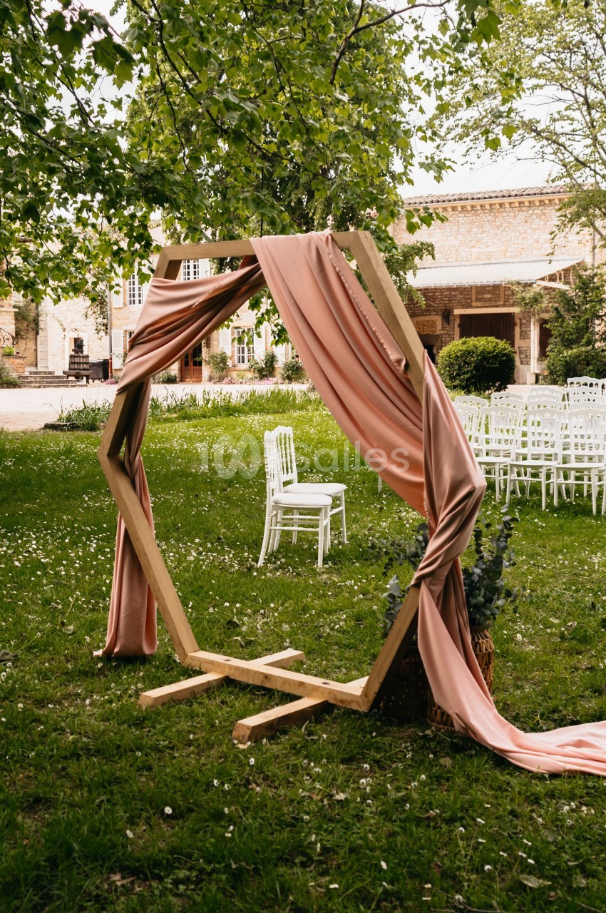 Arche hexagonale en bois décorée de drapés roses, installée sur une pelouse devant des chaises blanches et un bâtiment en…