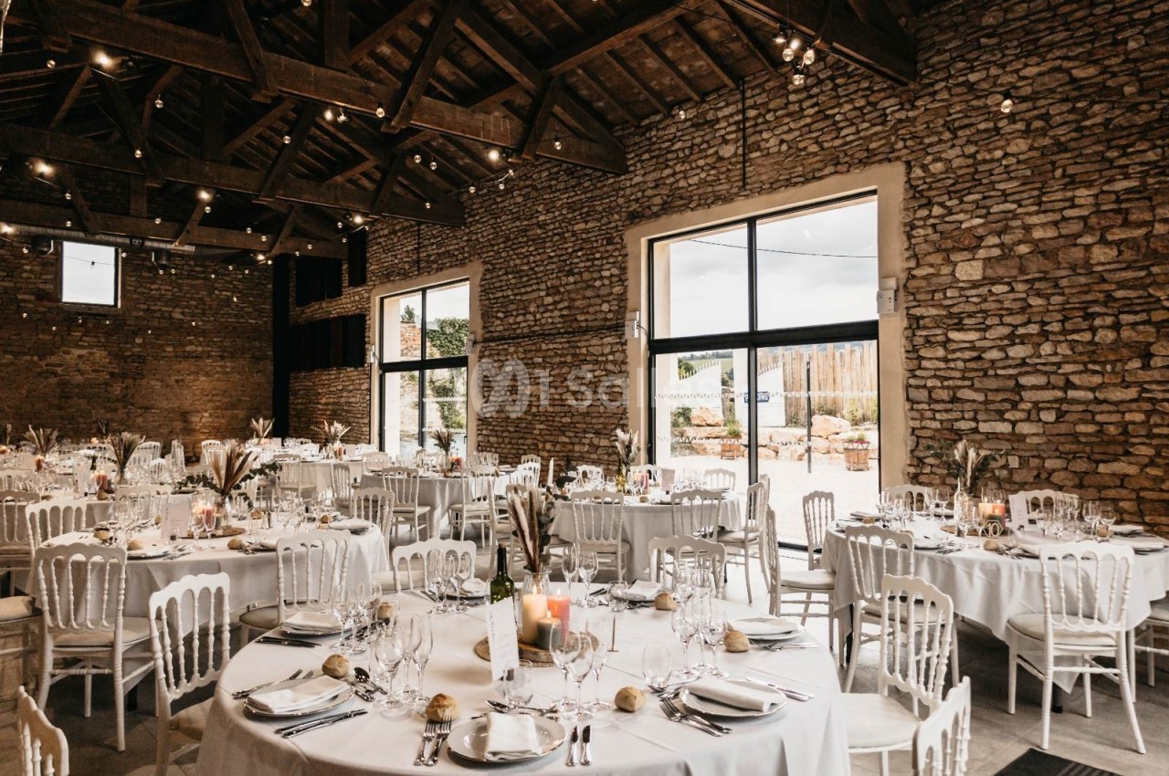 Salle de réception en pierre avec tables rondes dressées, chaises blanches et lumière naturelle entrant par de grandes baies…