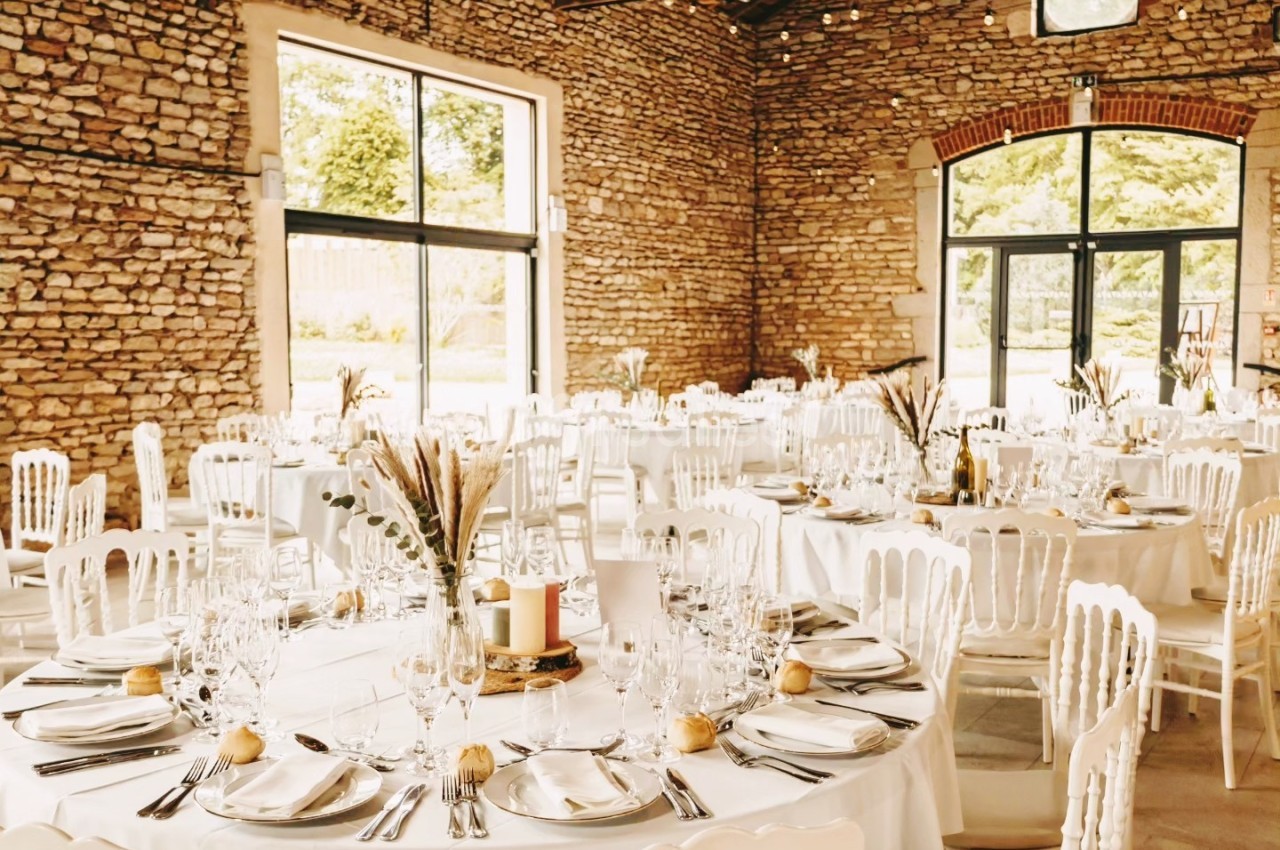 Salle de réception en pierres apparentes avec tables rondes dressées, décorées de bouquets et chaises blanches.