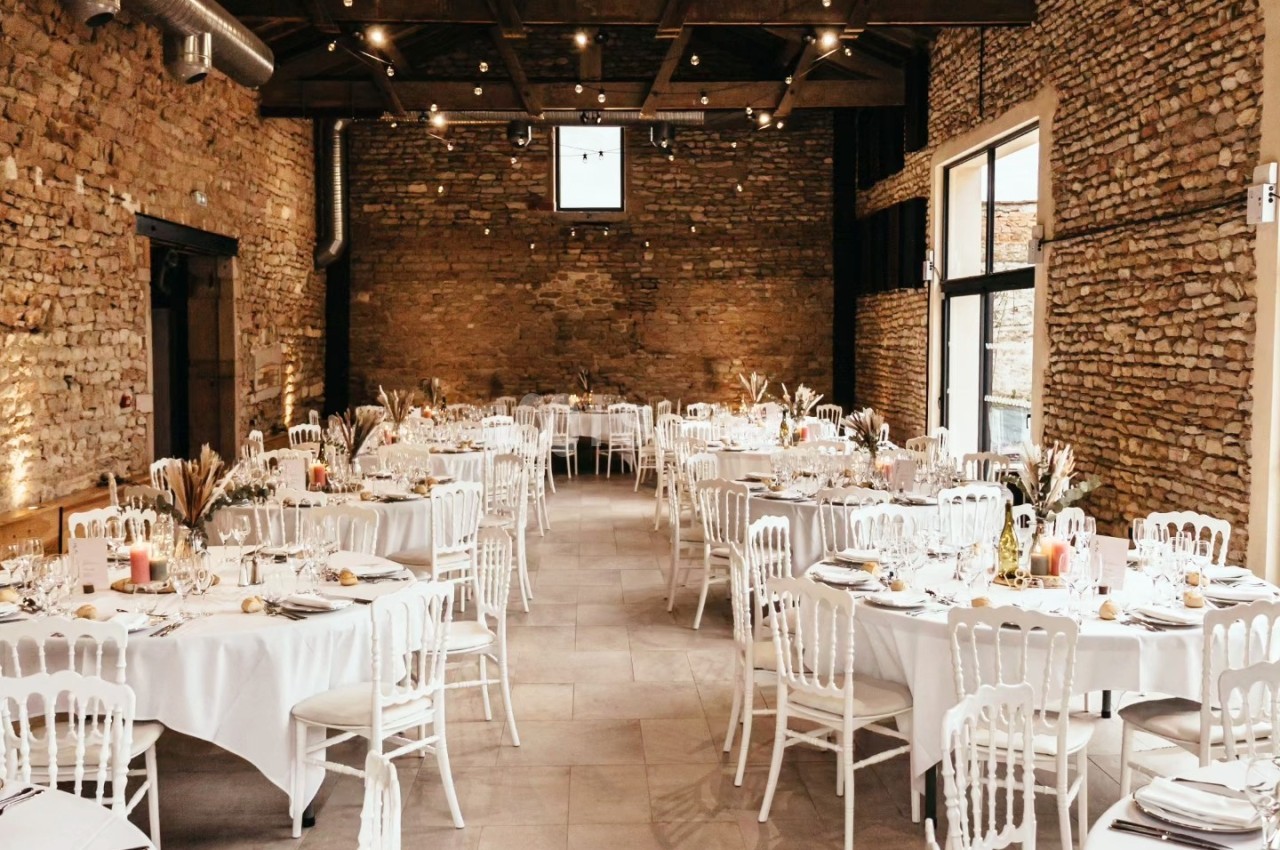Salle en pierre avec plafond en bois, tables rondes dressées avec nappes blanches et chaises blanches, éclairage chaleureux.