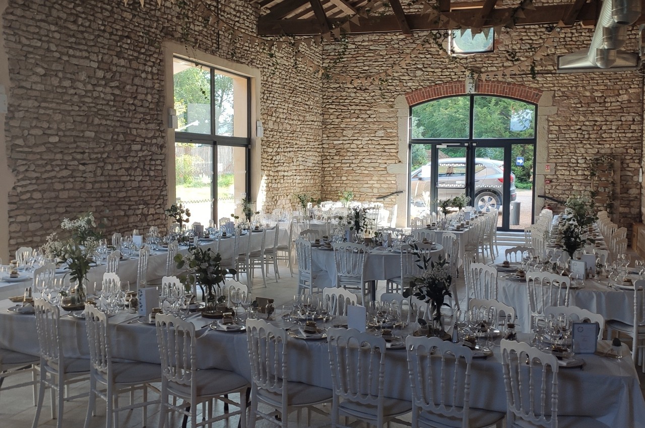 Salle de réception en pierre avec de longues tables décorées pour un événement, éclairée par de grandes baies vitrées.