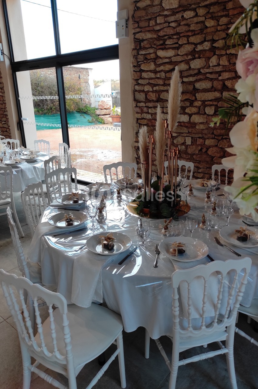 Salle de réception avec tables rondes dressées, nappes blanches, chaises blanches et décoration florale élégante.