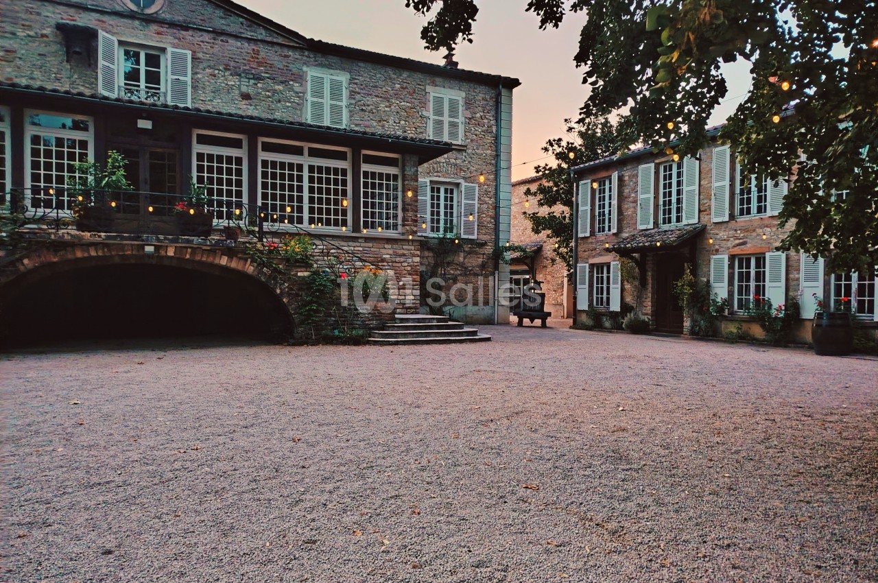 Cour pavée entourée de deux bâtiments en pierre avec volets blancs, éclairée par une lumière douce au crépuscule.