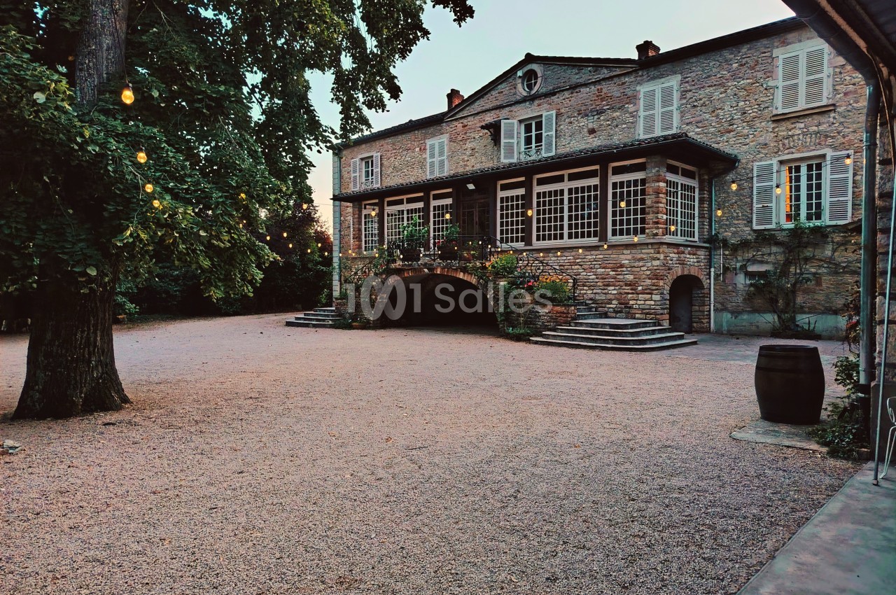 Cour d'une maison en pierre avec terrasse surélevée, entourée d'arbres et éclairée par des guirlandes lumineuses.