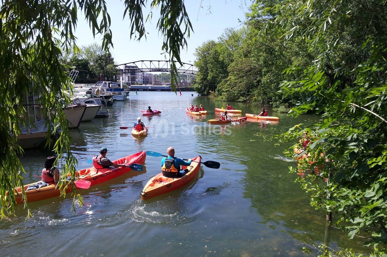 Des personnes pagayant en kayak sur une rivière bordée de végétation, avec des bateaux amarrés et un pont en arrière-plan.