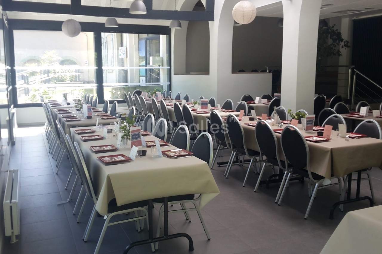 Salle lumineuse avec de longues tables dressées pour un repas, chaises alignées et décoration sobre.