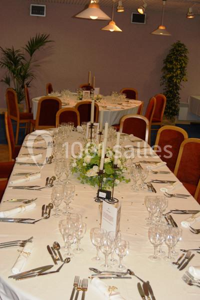 Salle de réception avec tables dressées, nappes blanches, couverts alignés, chandeliers et décoration florale centrale.