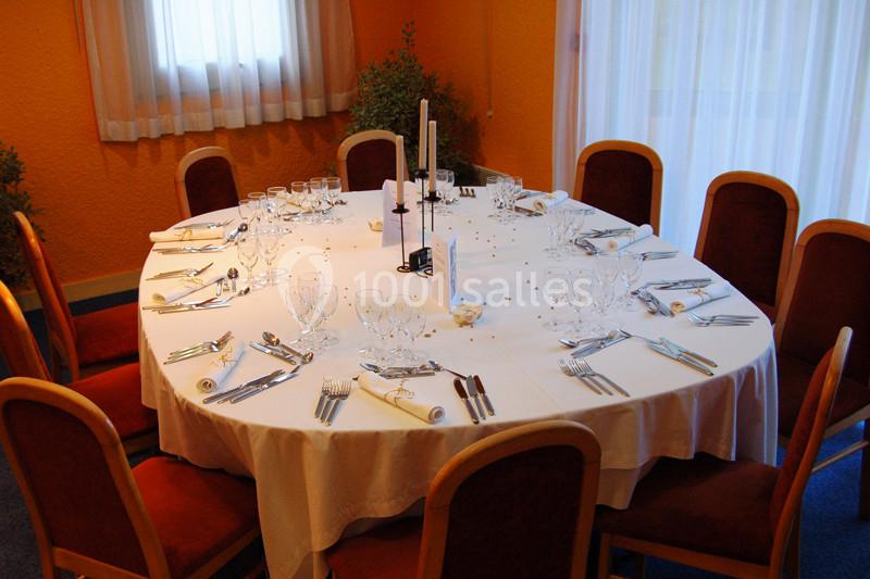 Table ronde dressée pour un repas, avec nappes blanches, couverts, verres et bougies, entourée de chaises en bois.