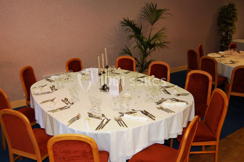 Table ronde dressée avec nappes blanches, verres, couverts et chandeliers, entourée de chaises en bois avec assise rouge.