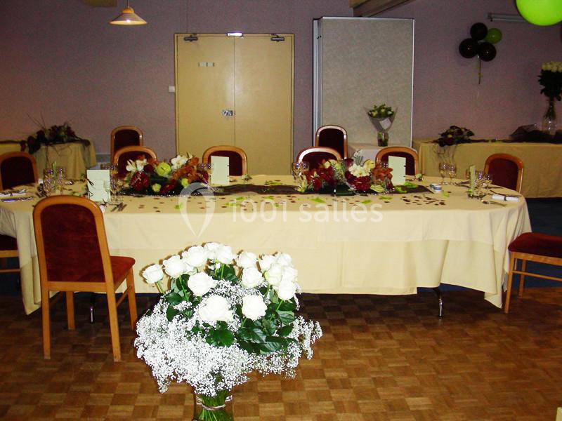 Salle décorée pour un événement avec une grande table ornée de fleurs, chaises alignées et bouquet de roses blanches au…