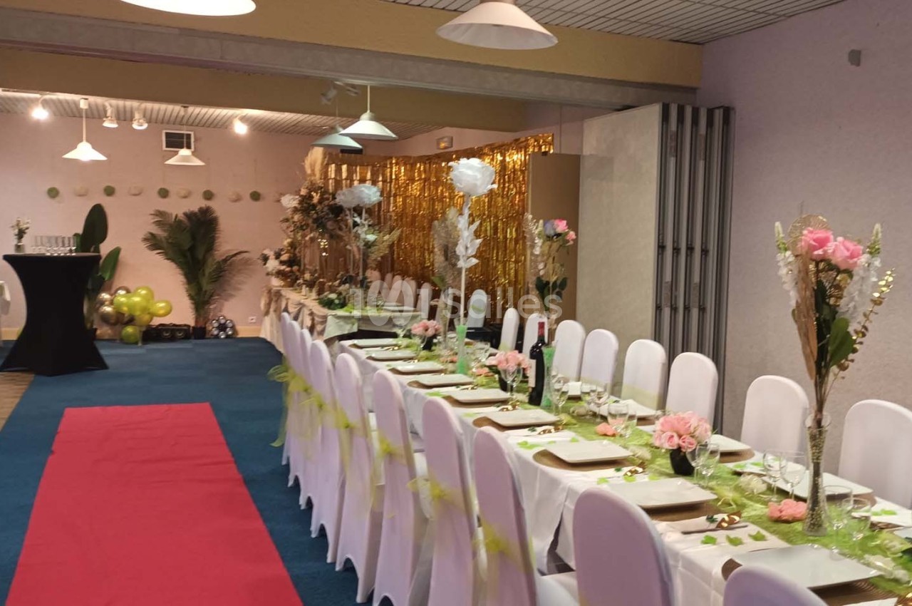 Salle décorée pour un événement avec une longue table dressée, chaises blanches, fleurs et tapis rouge au sol.