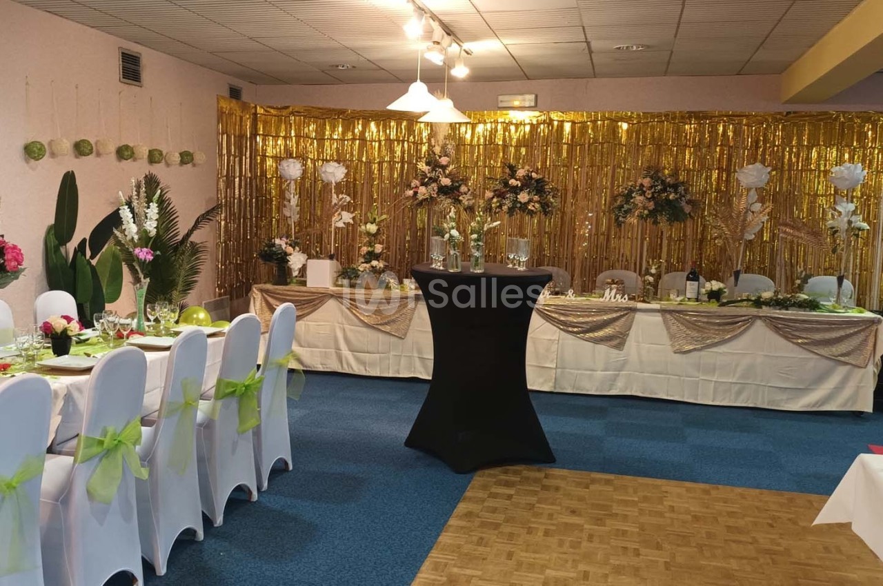 Salle décorée pour une réception avec tables dressées, chaises blanches ornées de nœuds verts et fond doré fleuri.