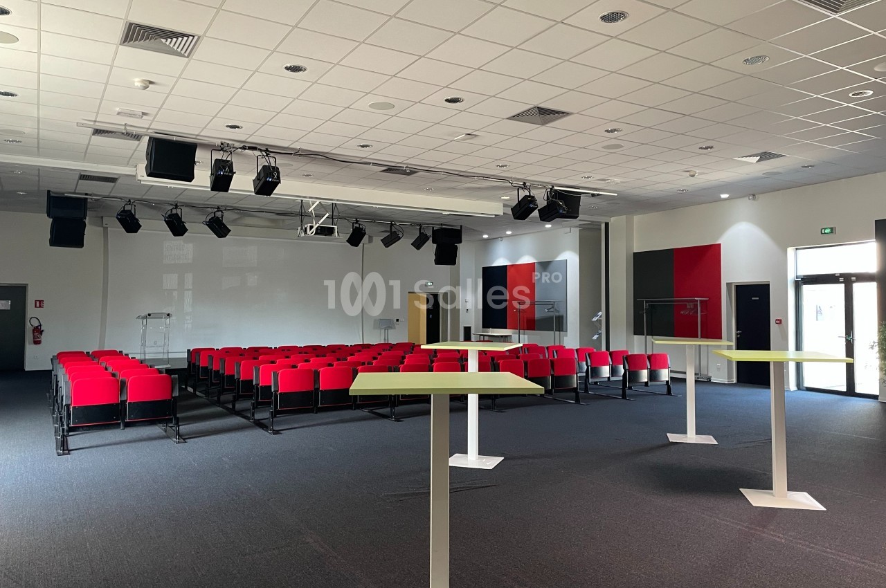 Salle de conférence moderne avec rangées de sièges rouges, pupitres blancs et éclairage suspendu.