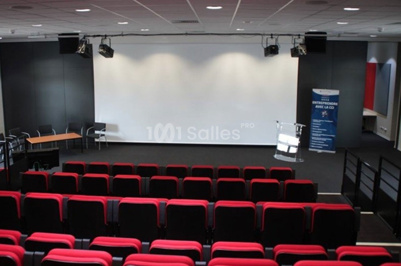 Salle de conférence vide avec rangées de sièges rouges, écran blanc, pupitre transparent et éclairage au plafond.