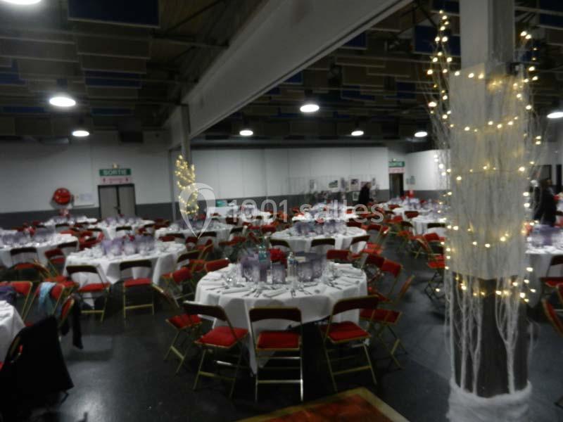 Salle décorée pour un événement, avec des tables rondes dressées, des chaises rouges et des lumières décoratives.