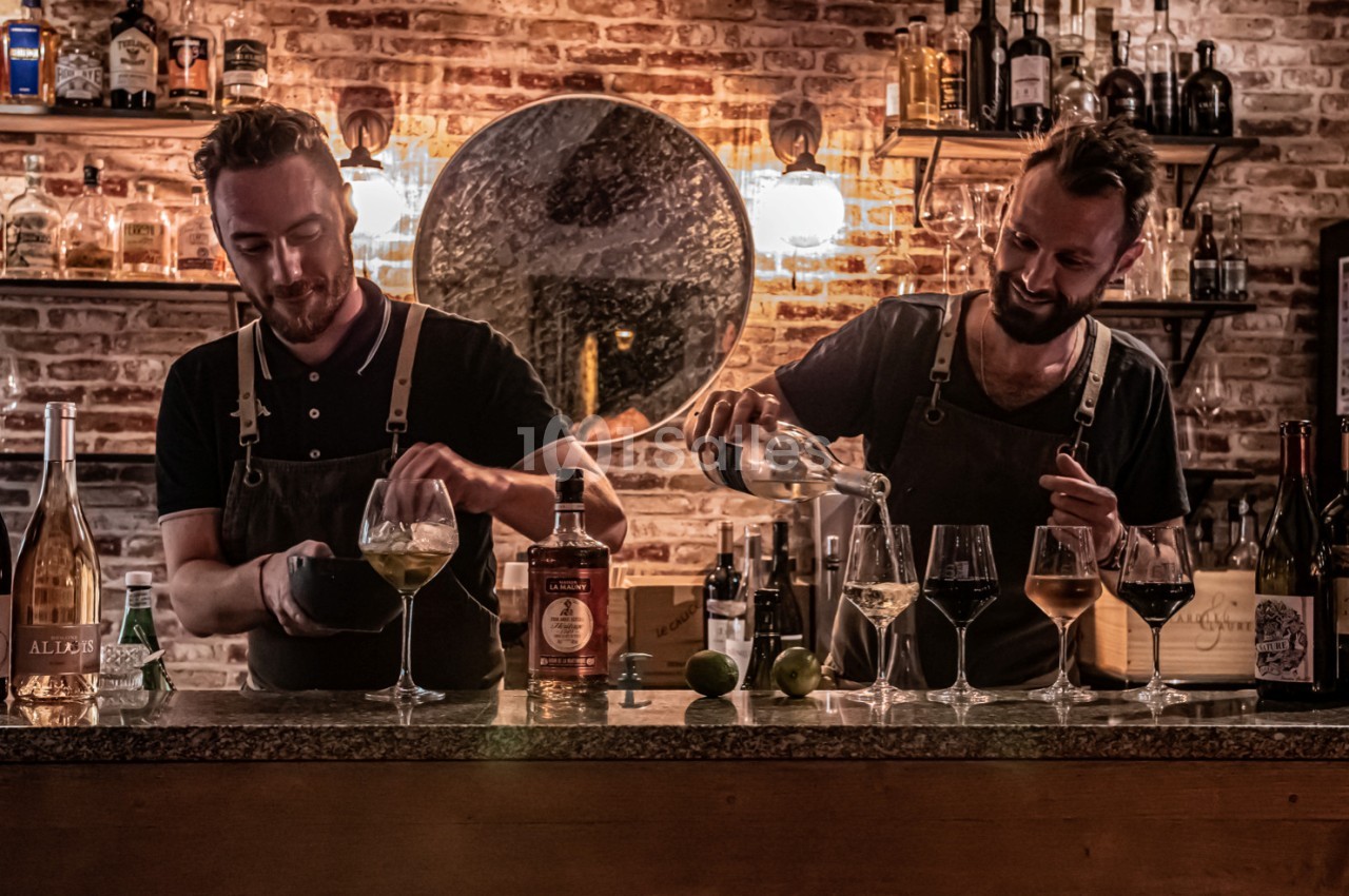 Deux barmans préparent des cocktails derrière un comptoir en bois, avec des bouteilles et des verres alignés.