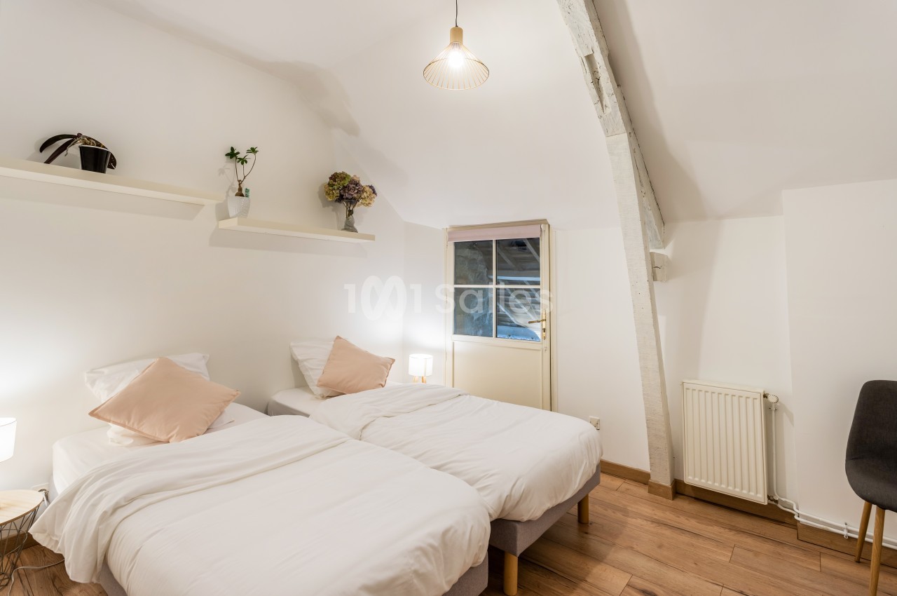 Deux lits simples avec draps blancs dans une chambre lumineuse, décorée de plantes et d'une fenêtre en arrière-plan.