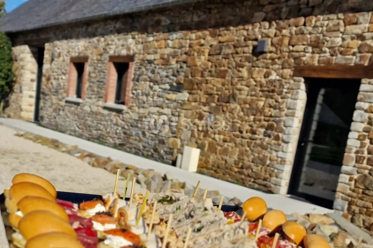 Plateau de bouchées apéritives variées devant un bâtiment en pierre avec toit en ardoise, sous un ciel bleu.