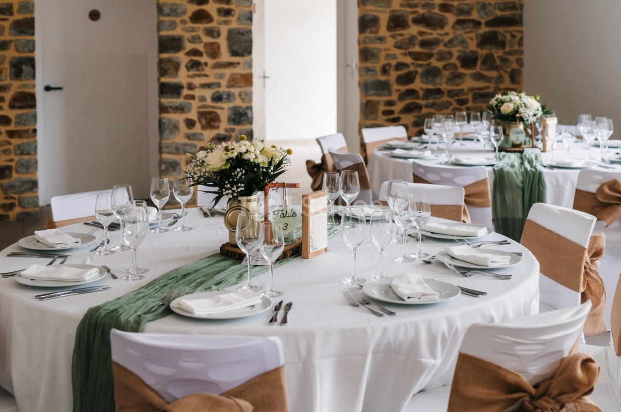 Salle décorée pour un événement avec tables rondes dressées, nappes blanches, chemins de table verts et chaises ornées de…