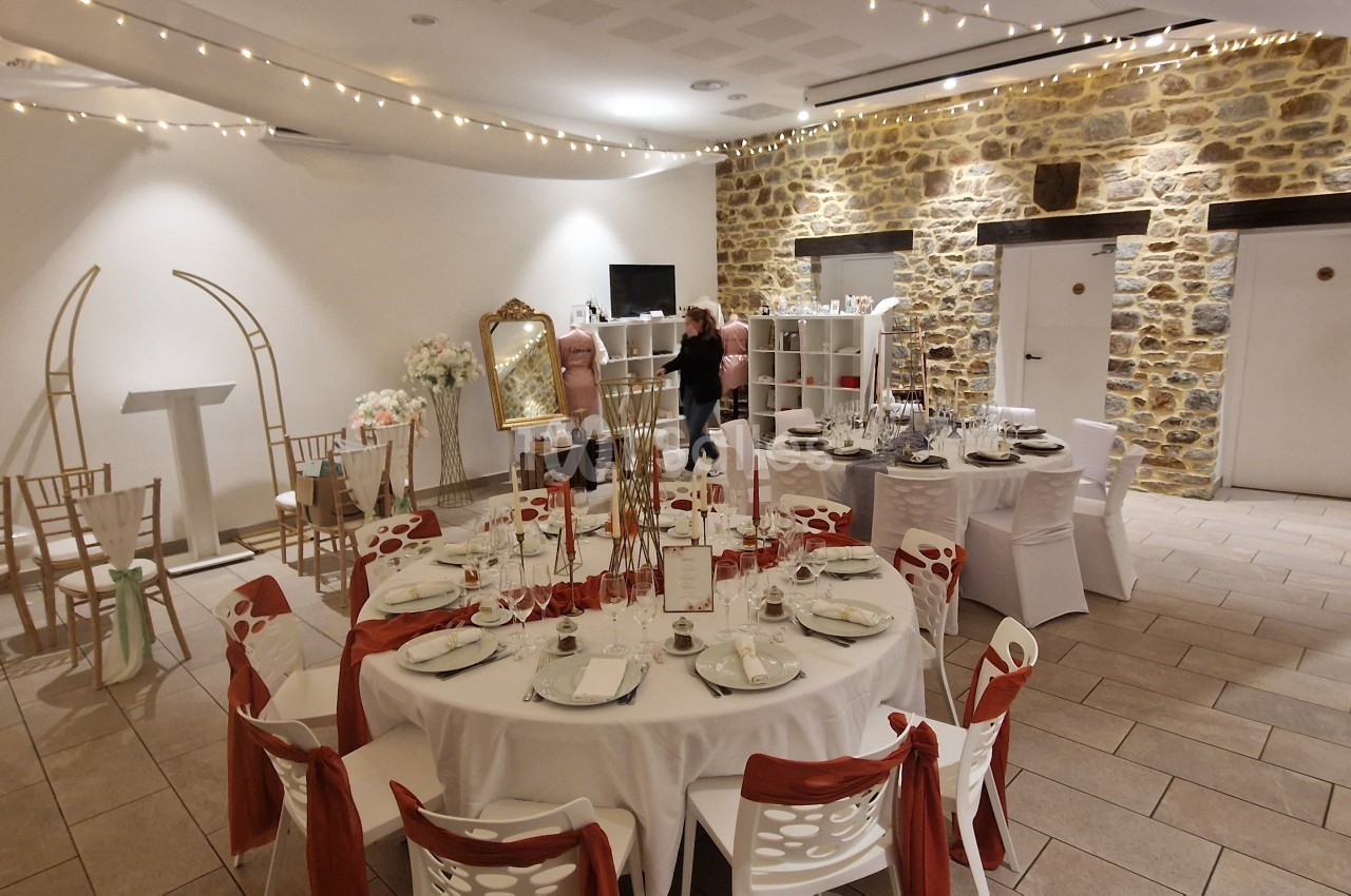 Salle de réception décorée avec des tables rondes, nappes blanches, chaises ornées de rubans rouges et guirlandes lumineuses.