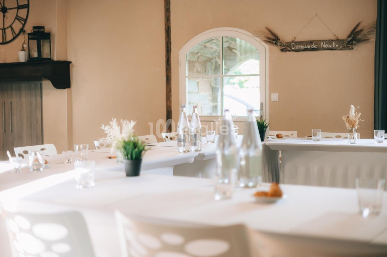 Salle lumineuse avec tables blanches décorées de plantes et bouteilles, près d'une fenêtre et d'un panneau mural.