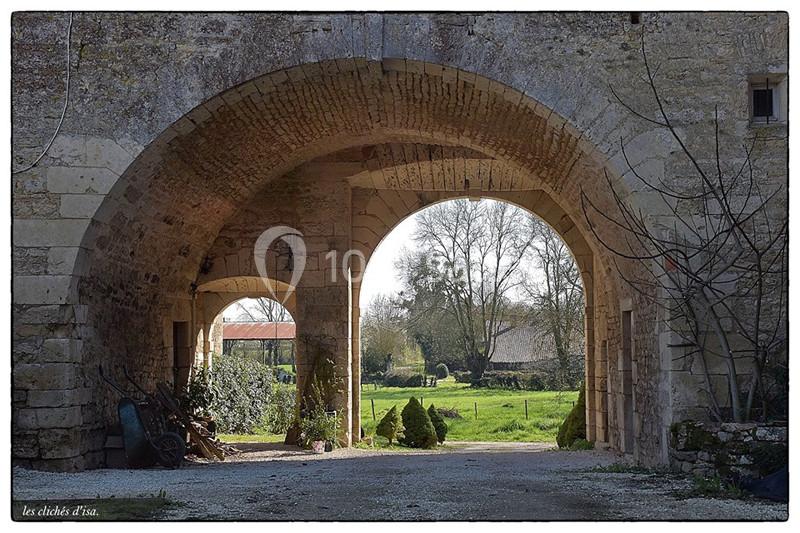 Vue d'une arche en pierre donnant sur un jardin verdoyant avec des arbres et des bâtiments en arrière-plan.