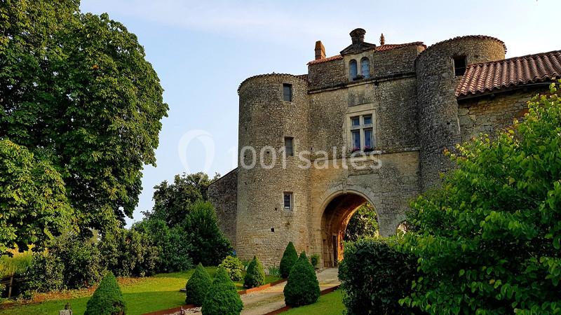 Château en pierre avec deux tours rondes, une arche centrale et un jardin verdoyant au premier plan.