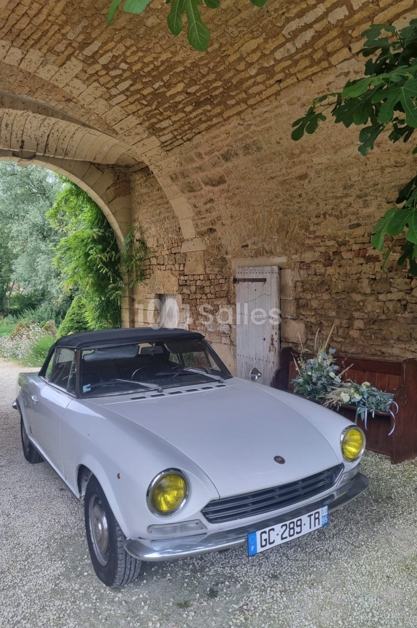 Voiture ancienne blanche décapotable garée sous une arche en pierre, entourée de végétation et de décorations florales.