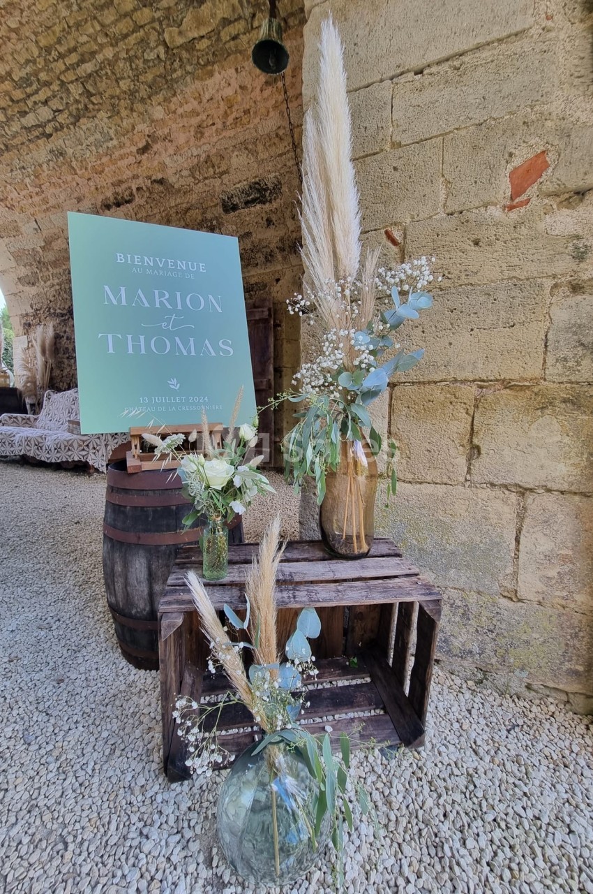 Décoration de mariage avec fleurs séchées, panneaux en bois, et affiche personnalisée sous une arche en pierre.