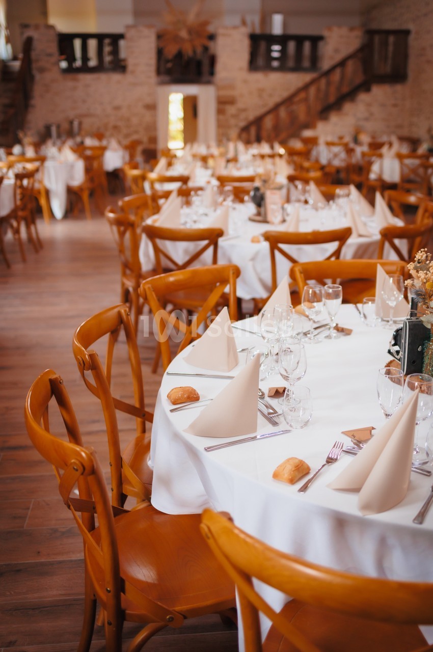 Salle de réception avec tables rondes dressées, nappes blanches, serviettes pliées et chaises en bois.