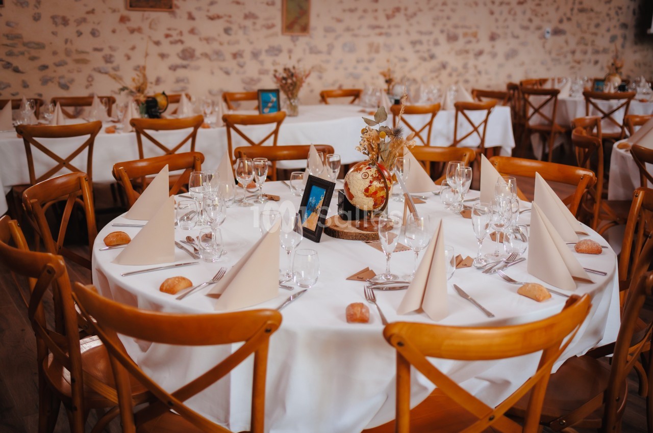 Salle de réception avec tables rondes dressées, nappes blanches, serviettes pliées, décorations florales et chaises en bois.