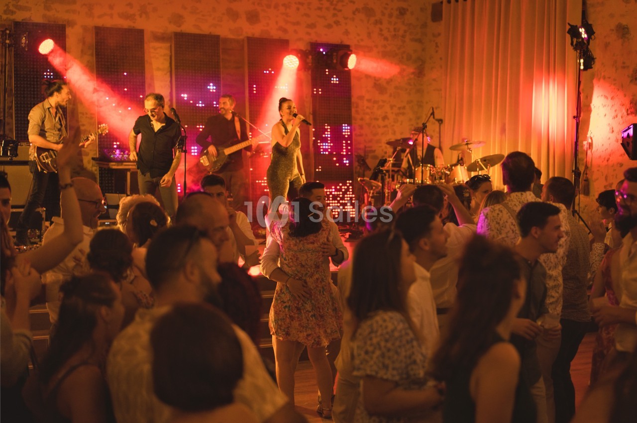 Groupe de musiciens jouant sur scène avec des lumières rouges, entourés d'invités dansant dans une salle en pierre.