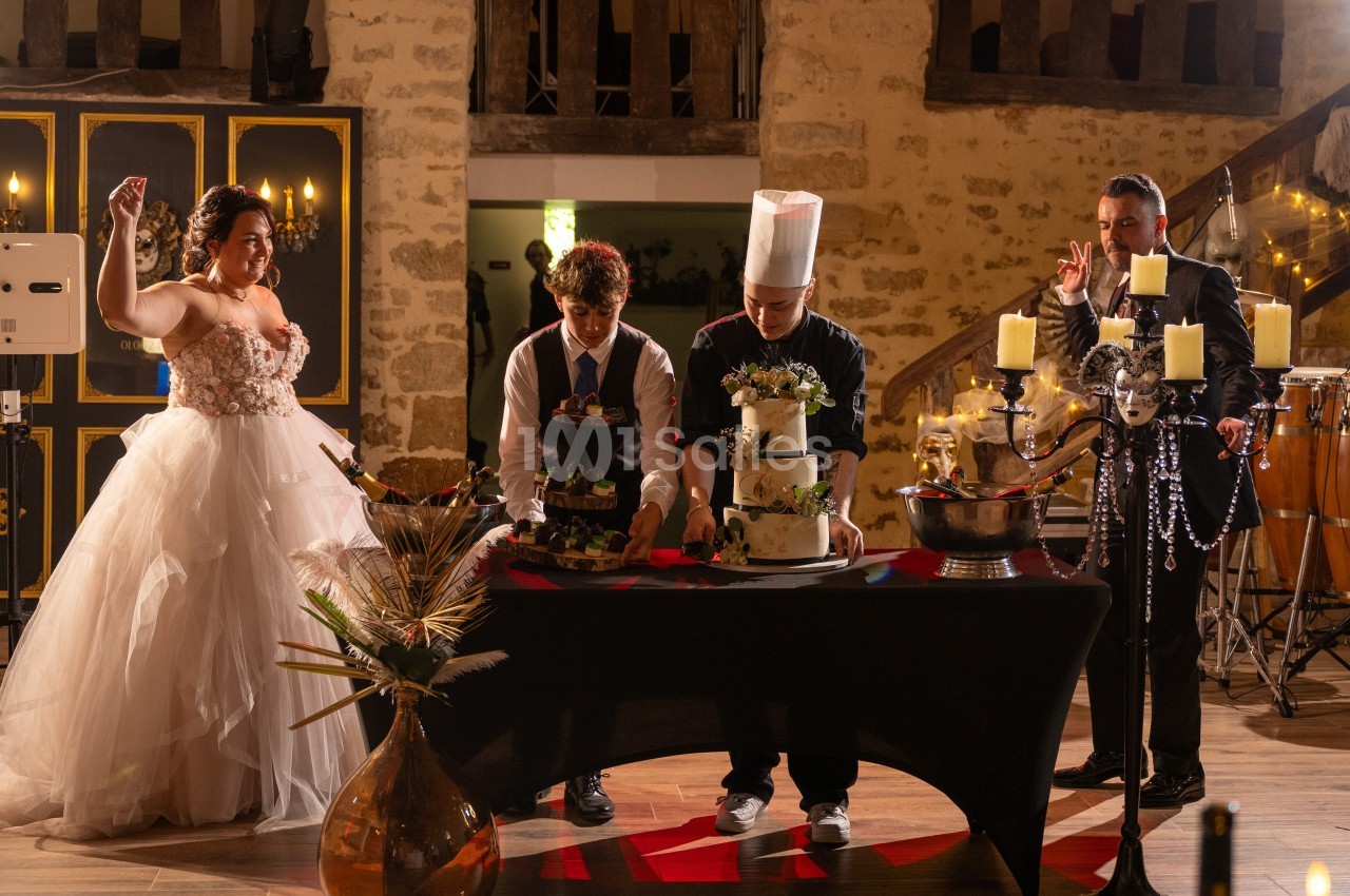 Un mariage avec une mariée en robe blanche, un serveur et un chef préparant un gâteau, dans une salle décorée.