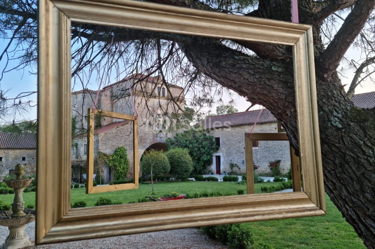 Cadres dorés suspendus à un arbre, encadrant une vue sur un jardin et un bâtiment en pierre.
