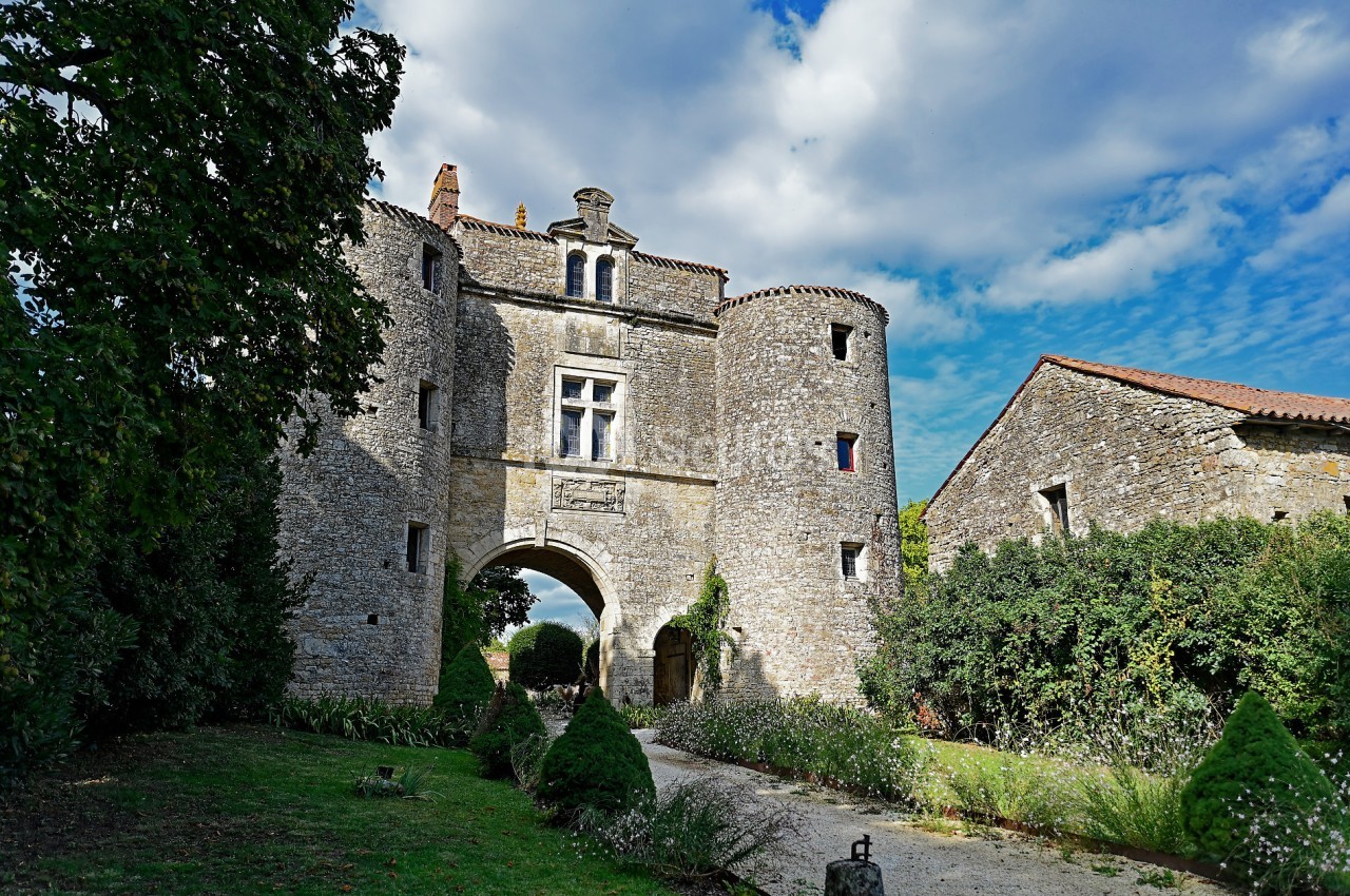 Entrée d'un château en pierre avec deux tours rondes, entourée de végétation sous un ciel partiellement nuageux.