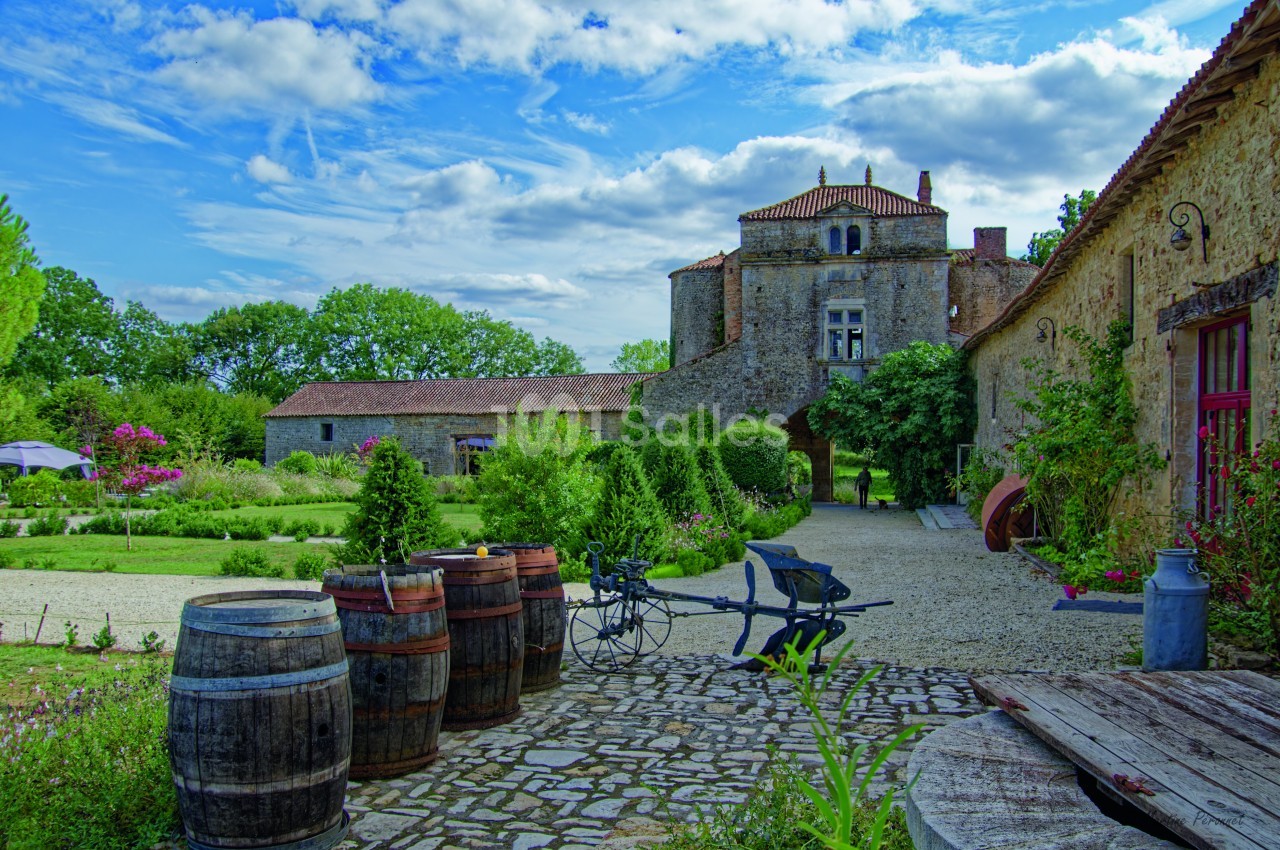 Cour pavée avec tonneaux, charrette et jardin fleuri, devant un château ancien sous un ciel partiellement nuageux.