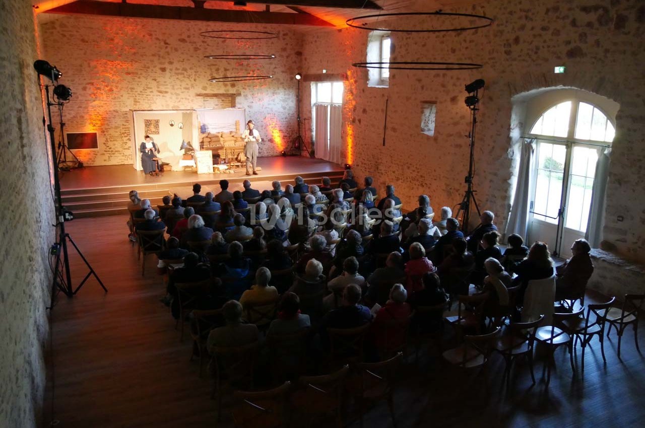 Salle en pierre avec un public assis face à une scène éclairée où se déroule une représentation théâtrale.