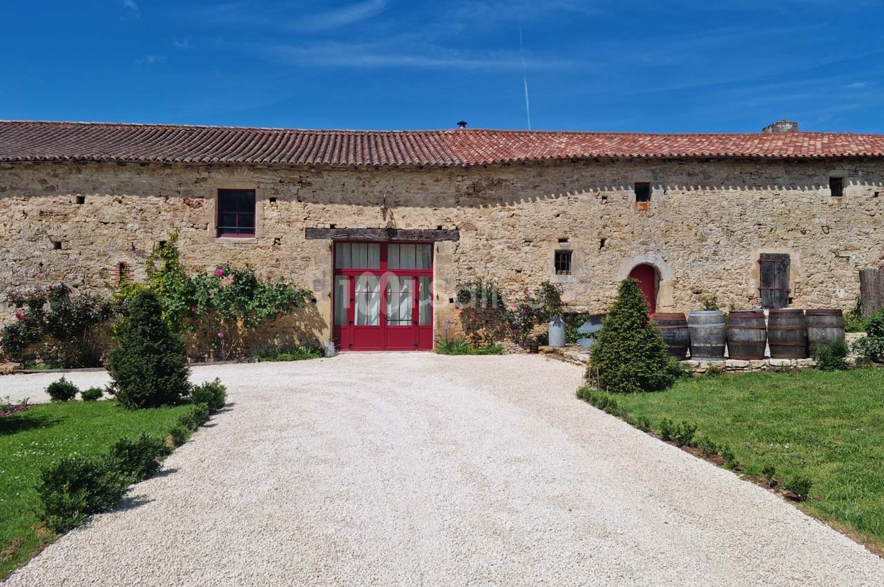 Façade en pierre d'un bâtiment ancien avec porte rouge, entourée de verdure et de barriques en bois.
