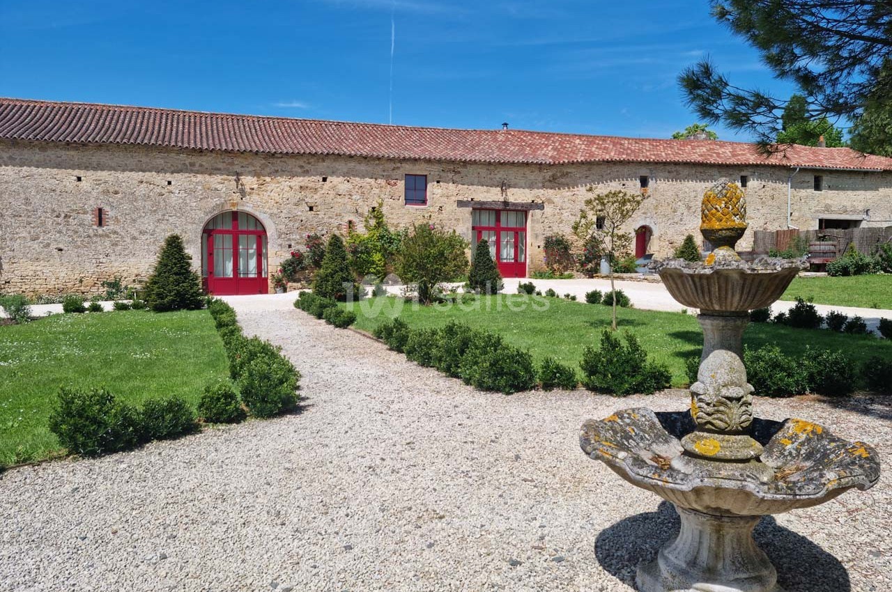 Cour d'une bâtisse en pierre avec une fontaine au premier plan, jardin aménagé et ciel bleu en arrière-plan.