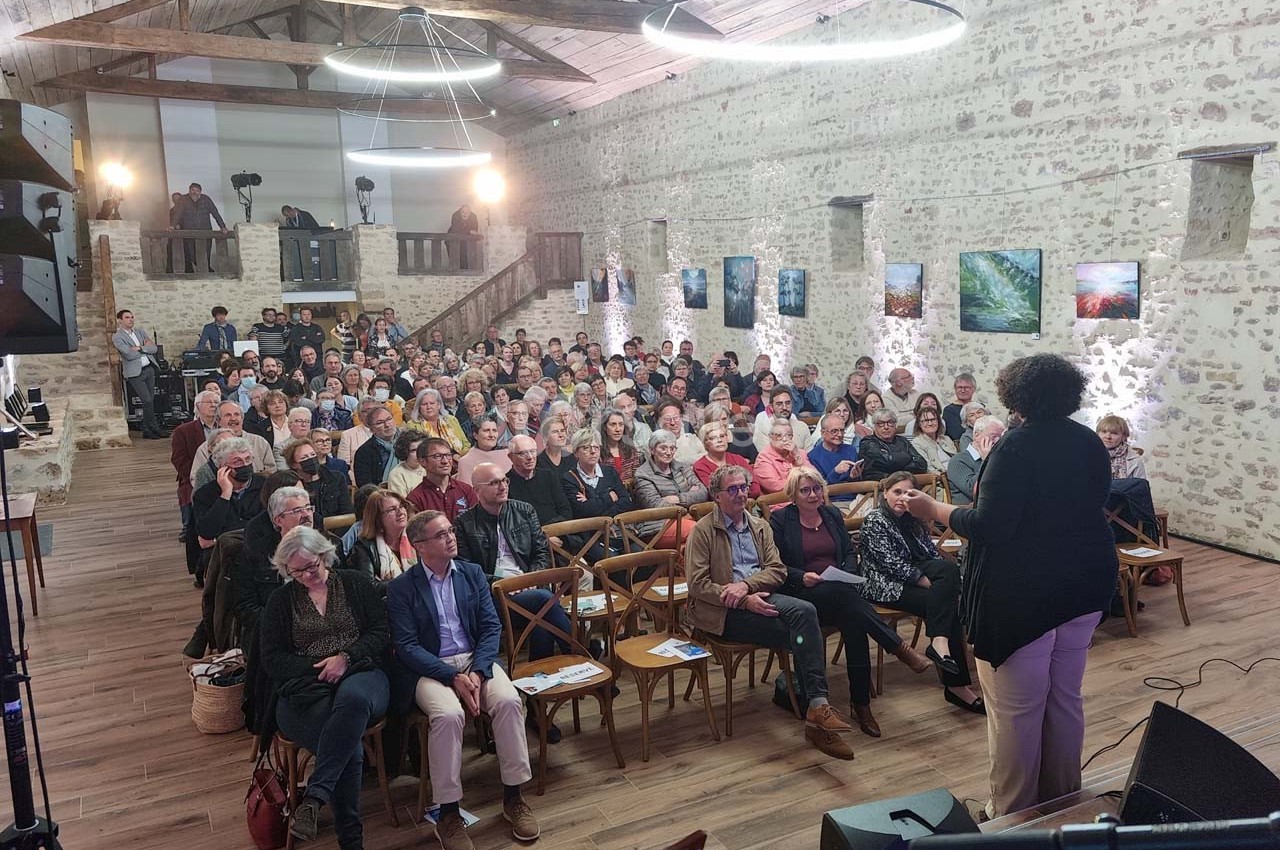 Une conférencière s'adresse à un public assis dans une salle en pierre avec des photos accrochées aux murs.