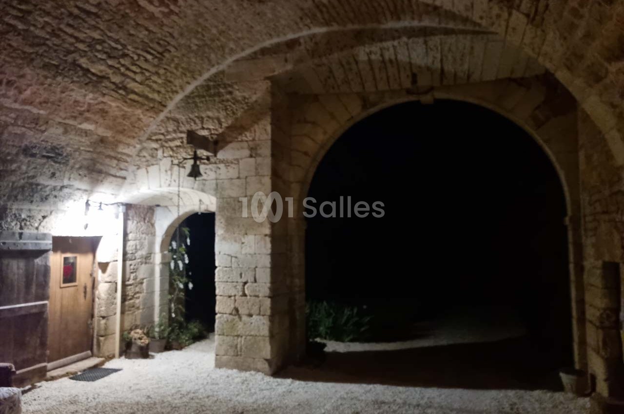 Entrée voûtée en pierre éclairée, avec une porte en bois à gauche et une ouverture sombre donnant sur l'extérieur.