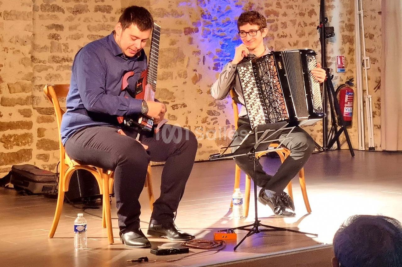 Deux musiciens assis jouent de la guitare électrique et de l'accordéon sur une scène éclairée dans une salle en pierre.