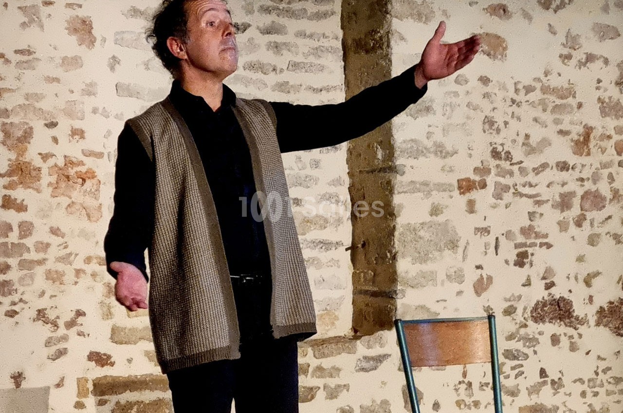 Un homme debout, bras ouverts, s'exprime devant un mur en pierre, avec une chaise en bois à côté de lui.