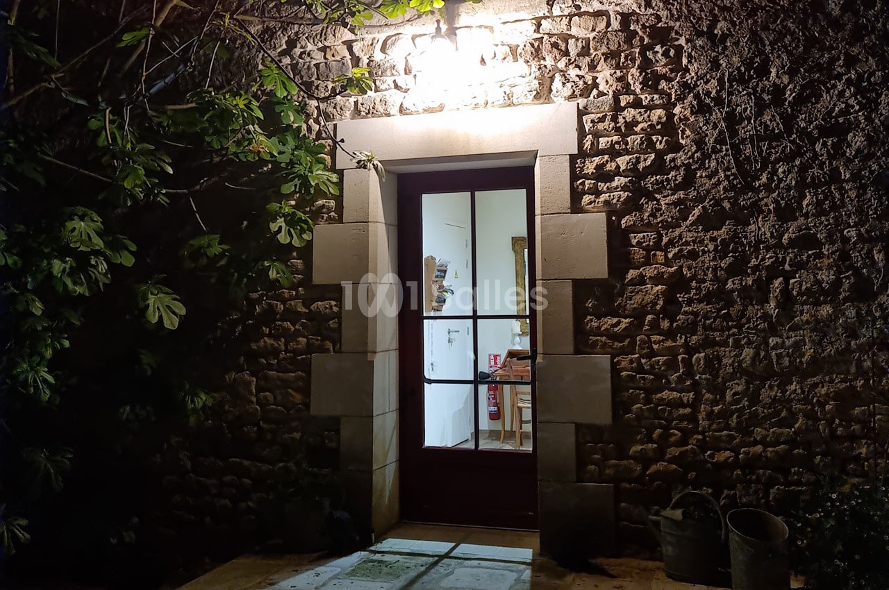 Porte vitrée éclairée donnant sur un intérieur, encadrée par un mur en pierre et entourée de végétation.