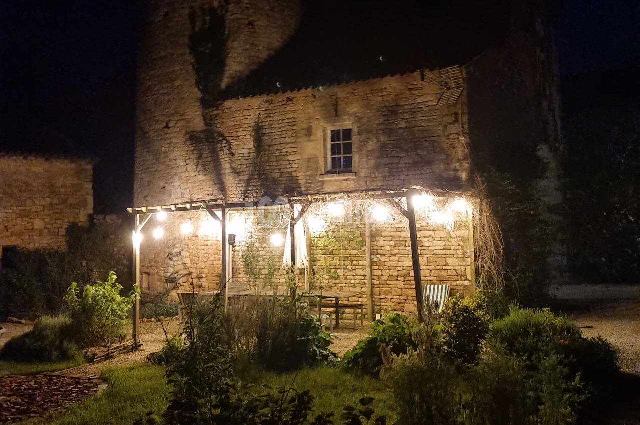 Pergola éclairée par des guirlandes lumineuses devant un bâtiment en pierre, entourée de végétation, de nuit.