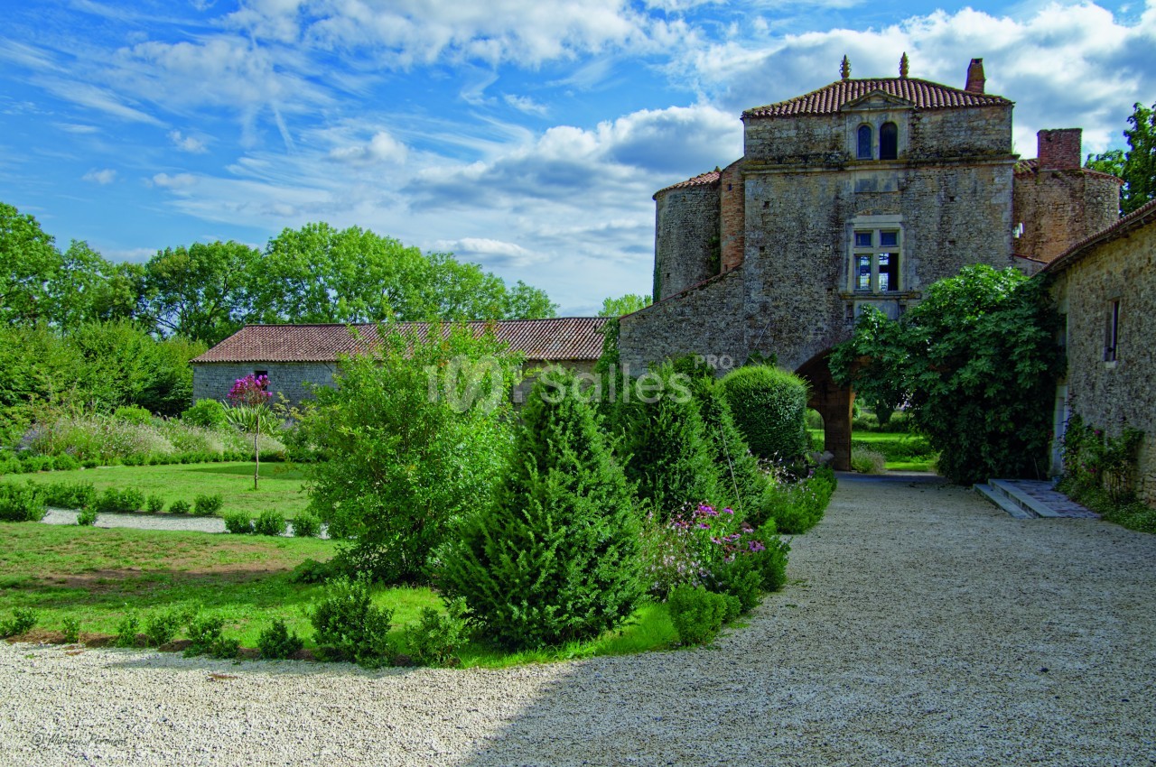 Allée gravillonnée menant à un bâtiment en pierre entouré de jardins verdoyants sous un ciel partiellement nuageux.