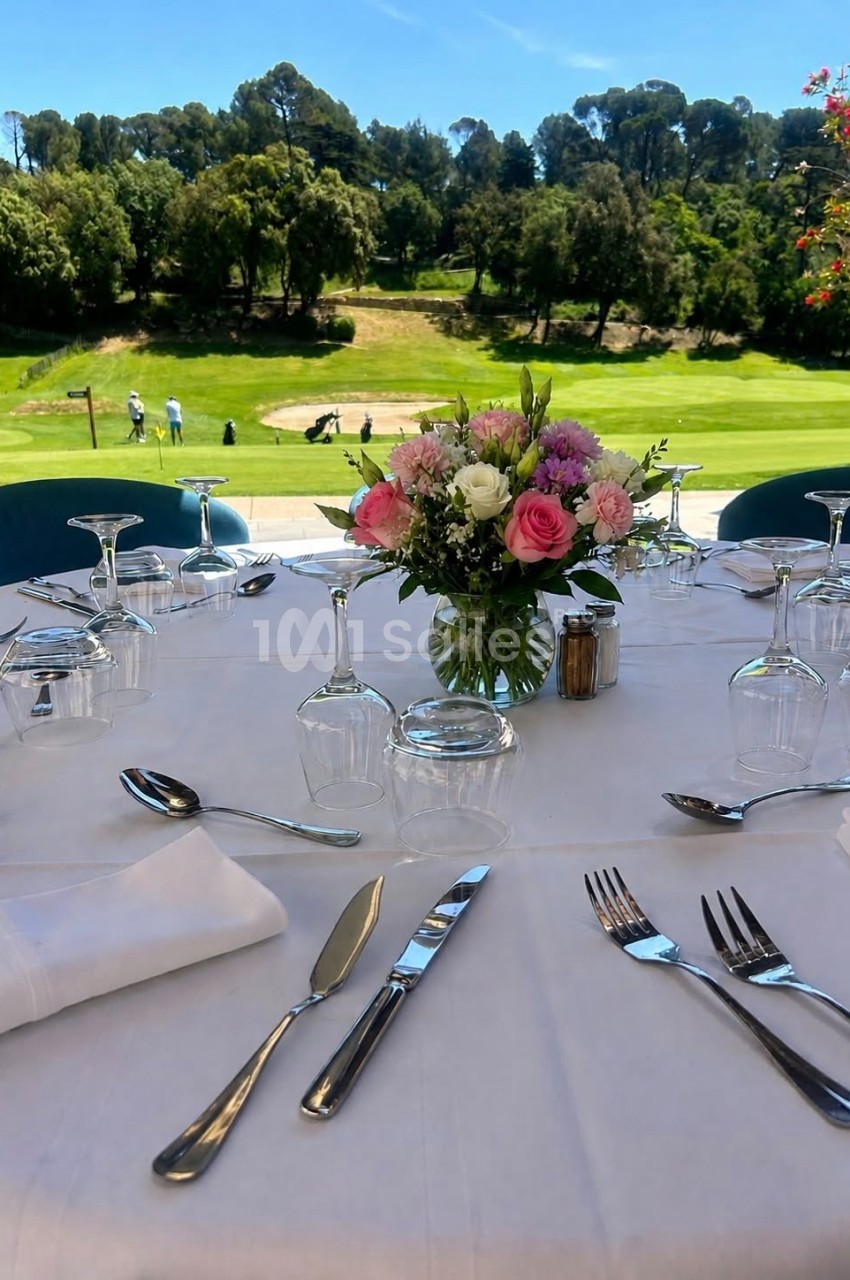 Table dressée avec nappes blanches, couverts et bouquet de fleurs, vue sur un terrain de golf en arrière-plan.