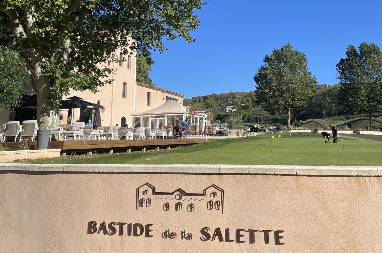 Terrasse d'un bâtiment beige avec des tables, vue sur un terrain de golf et un panneau ’Bastide de la Salette’.