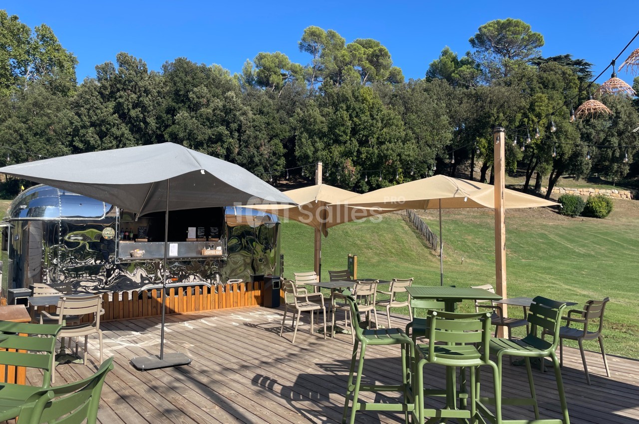Terrasse en bois avec tables, chaises et parasols, située près d'un food truck métallique dans un cadre verdoyant.